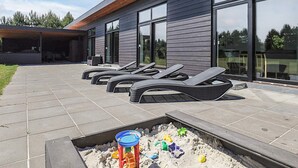 A modern building with large windows and a patio area with lounge chairs and a children's play area. - 5 Star Holiday Home in Vaeggerlose (Vaeggerlose)
