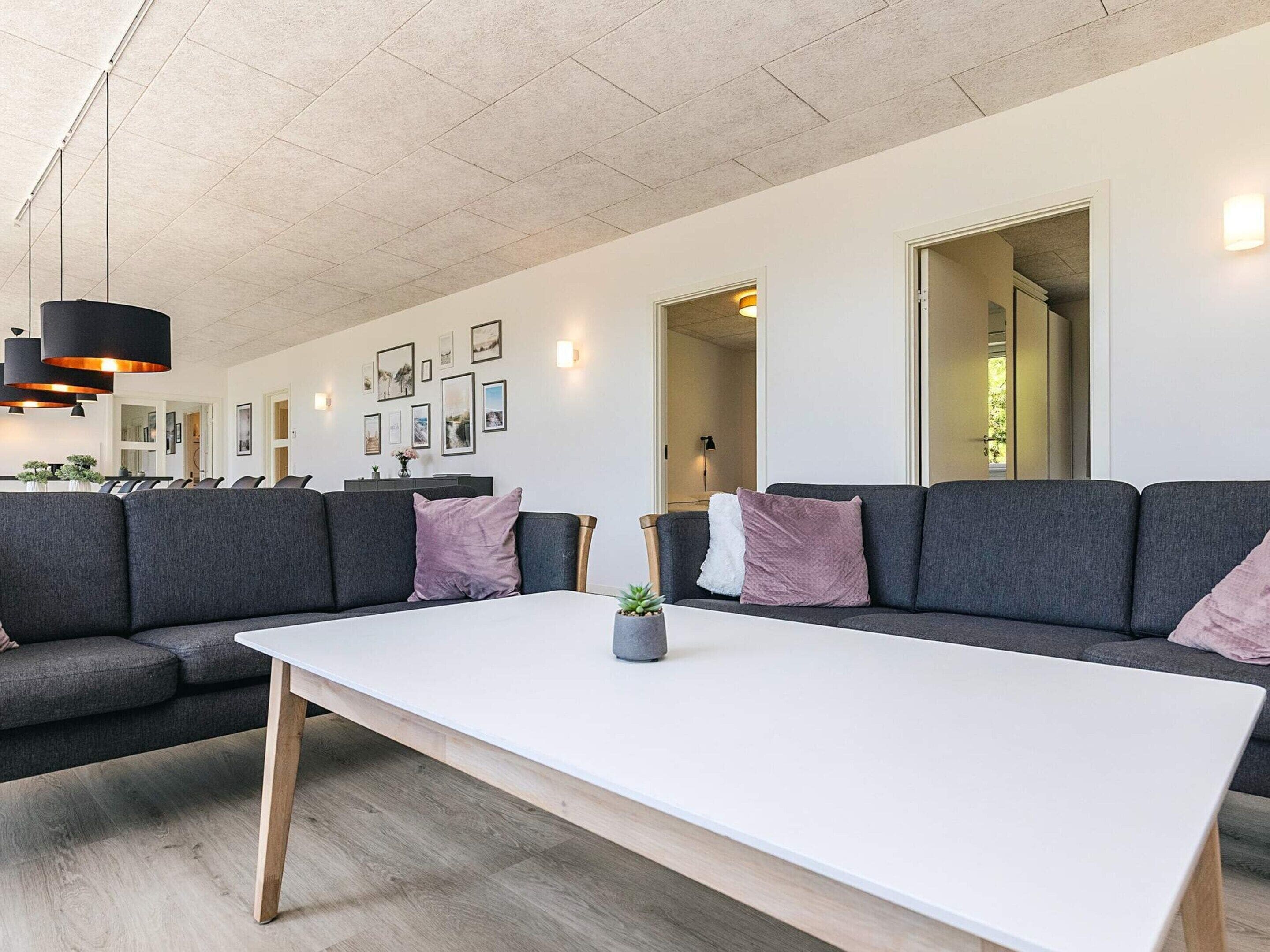 A modern living room with a large white table, grey sofas, and a dining area in the background.