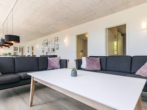 A modern living room with a large white table, grey sofas, and a dining area in the background.