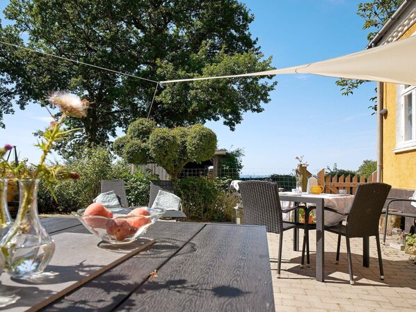 An outdoor dining area with a table and chairs, surrounded by greenery and a clear sky.