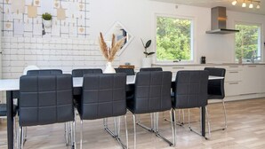 Et moderne køkken med spiseplads, komplet med et hvidt flisebeklædt backsplash, et stort vindue med udsigt over grøn vegetation, samt en central ø med en vase tørrede blomster.
