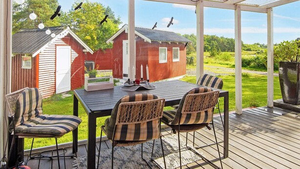 En overdækket terrasse med et bord og stole, der kigger ud over et rødt ladebygning og en græsplæne.