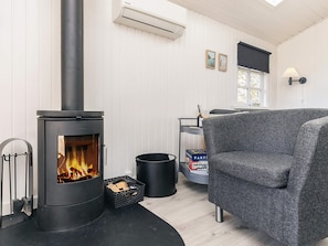A cozy room with a wood-burning stove, a grey upholstered chair, a small table with books, and a window with a view.
