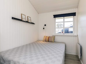 A small, single-bed room with a window, a wall-mounted shelf with pictures, and a bedside lamp.