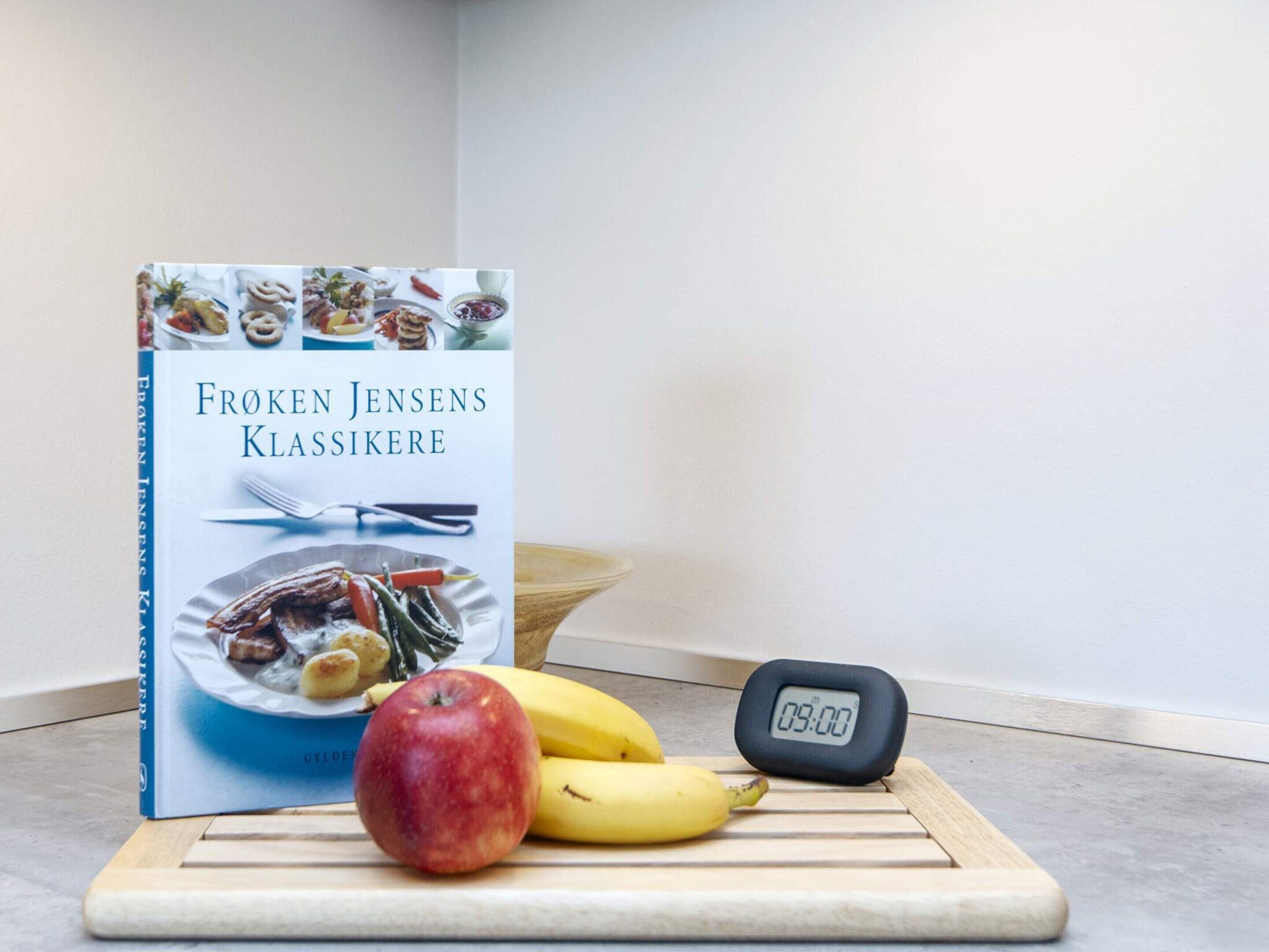 Un livre intitulé « FROKEN JENSENS KLASSIKERE » est posé sur une surface en bois, à côté d’une horloge numérique qui affiche 09 h 00, d’une pomme et de bananes.