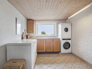 A compact kitchen with a built-in washer and dryer, wooden cabinets, a small stool, and a window providing natural light. - 8 Person Holiday Home in Veslos (Vesløs)