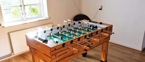 A foosball table in a room with wooden flooring, a dartboard on the wall, and a window providing natural light.
