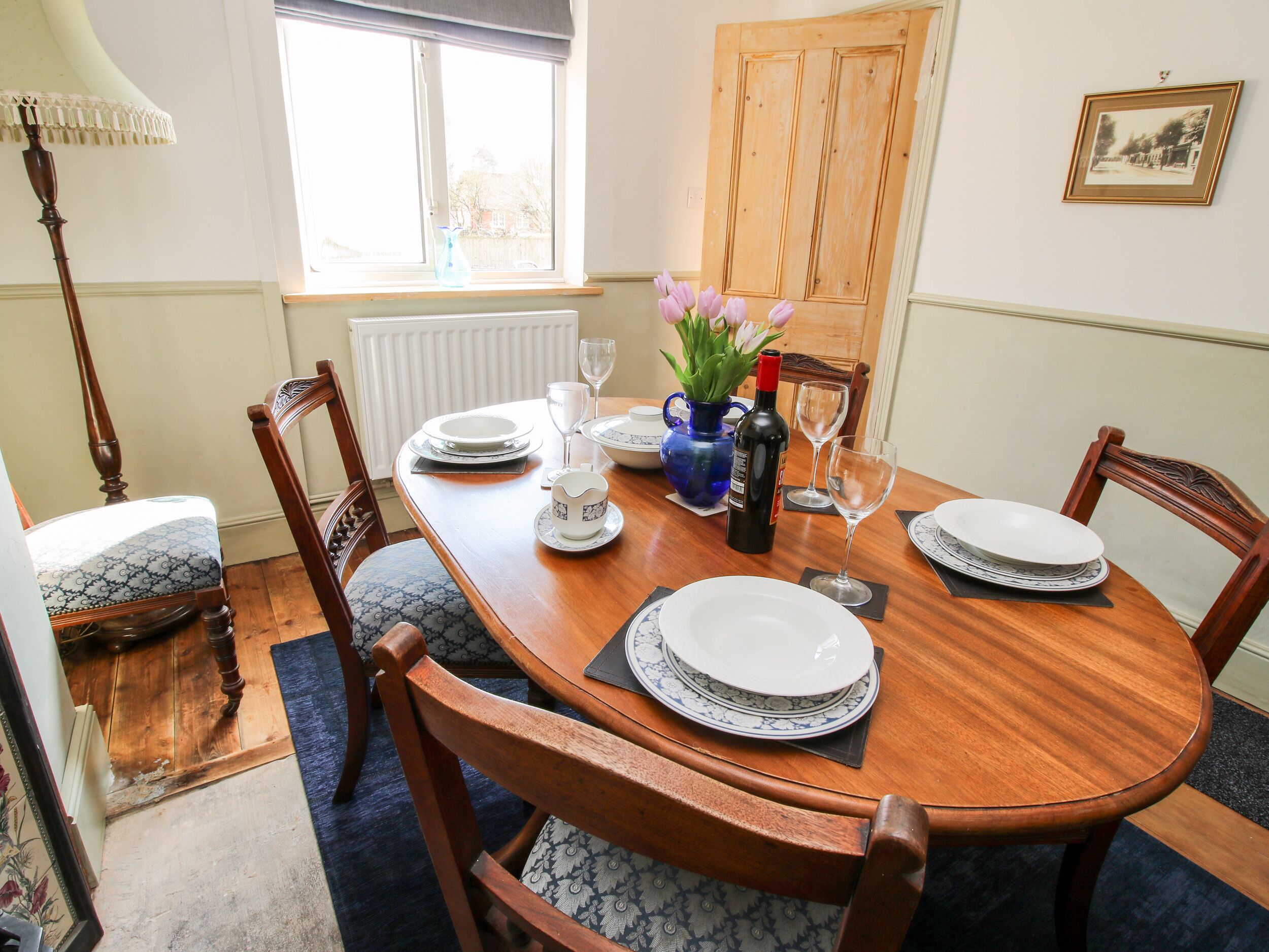 A dining room with a wooden table set for four, surrounded by chairs with patterned cushions. A vase with flowers and a bottle of wine are on the table.