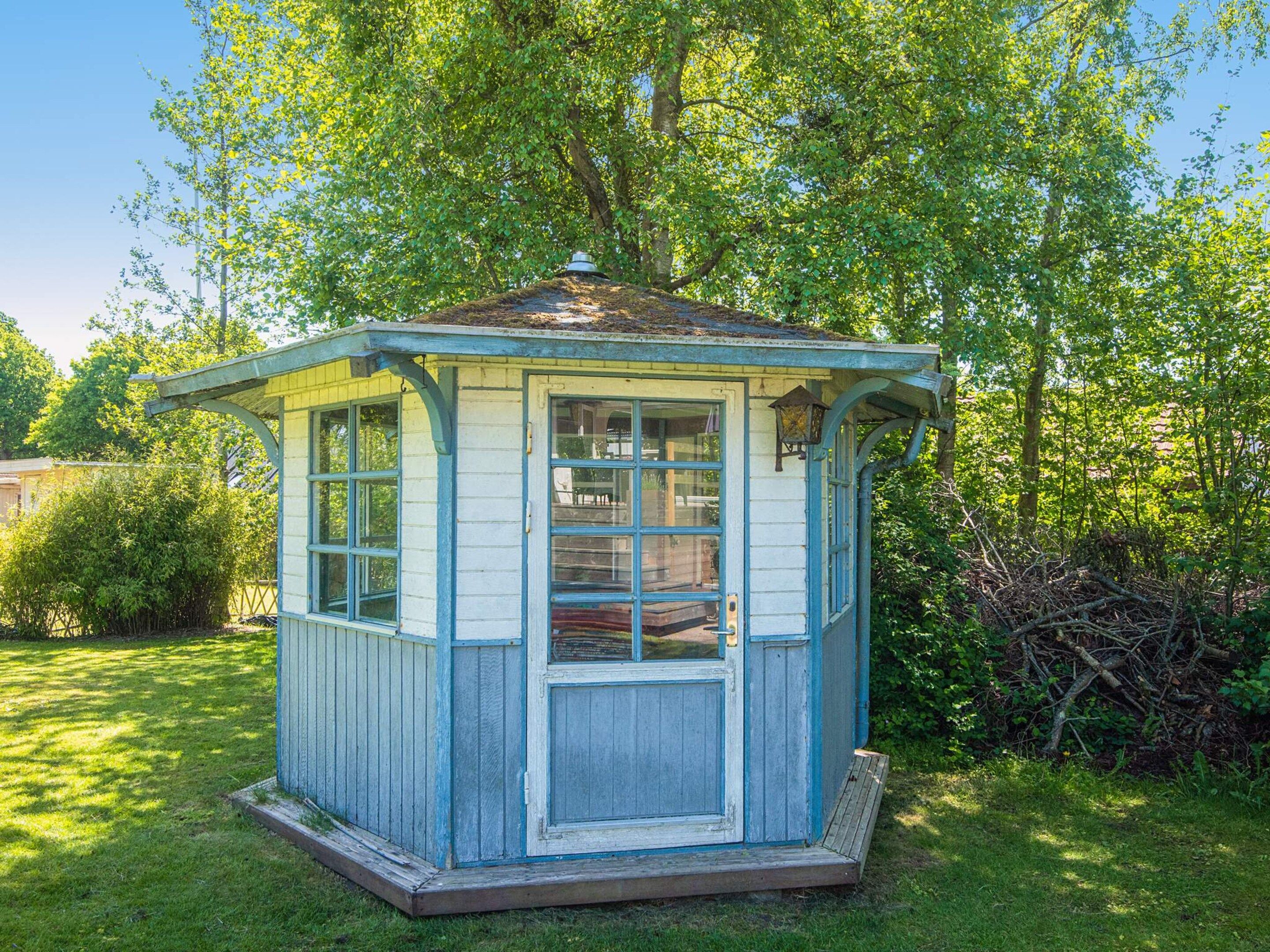 A small, single-story wooden structure with a peaked roof, a door, and a window, surrounded by greenery.