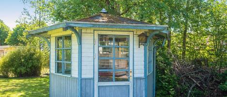 A small, single-story wooden structure with a peaked roof, a door, and a window, surrounded by greenery.