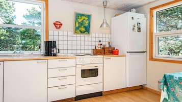 A compact kitchen with white cabinets, a stove, a refrigerator, and a window with a view of trees.