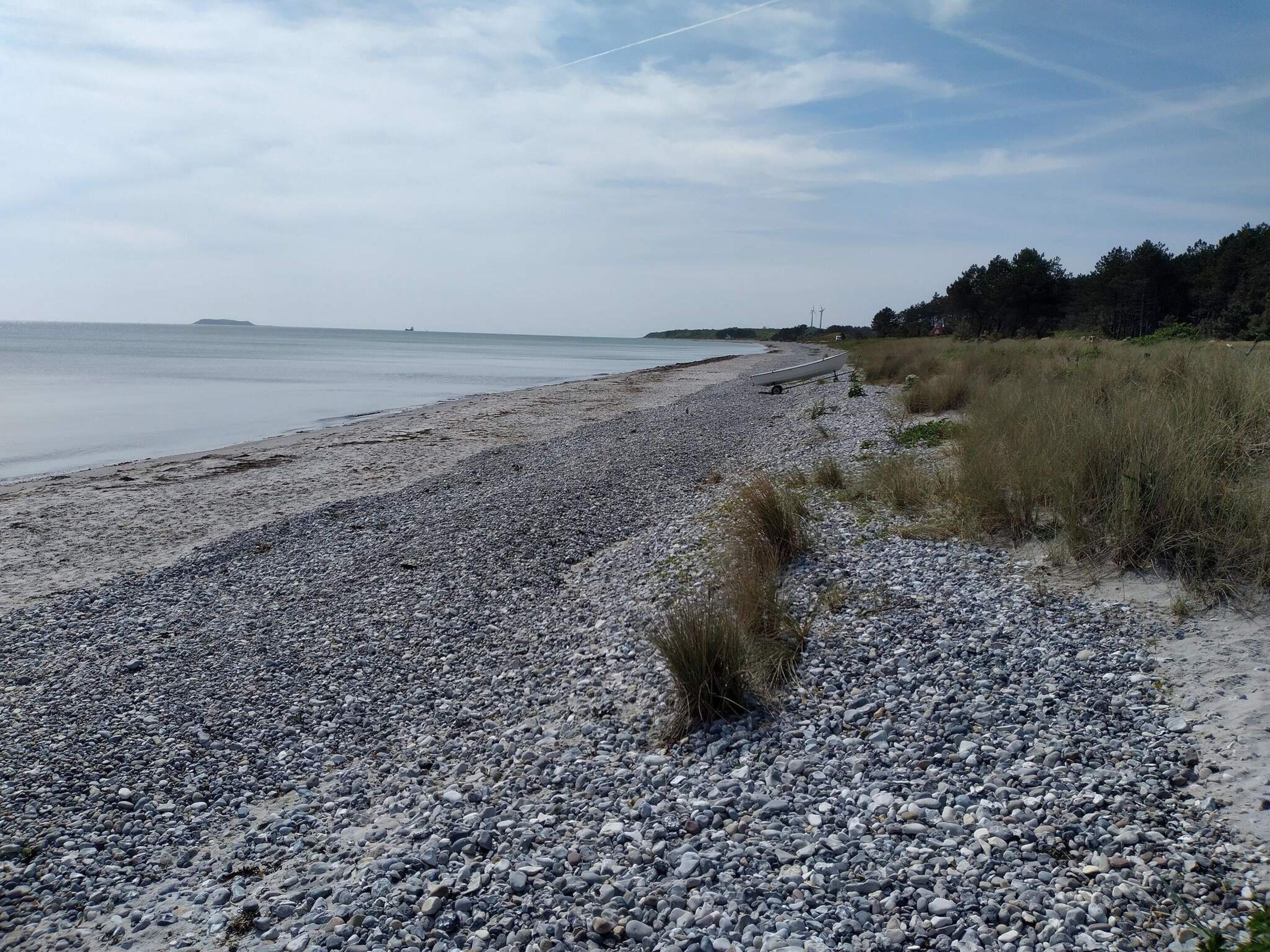 A pebbled beach with a boat, grassy dunes, and a distant shoreline.