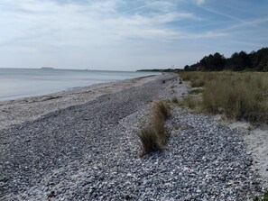 A pebbled beach with a boat, grassy dunes, and a distant shoreline. - 4 Person Holiday Home in Ebeltoft (Ebeltoft)