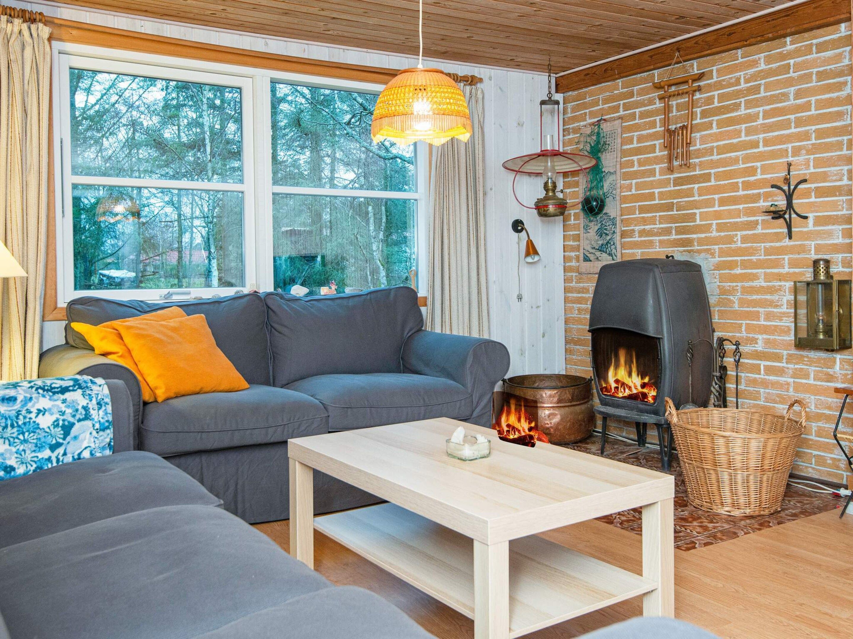 A cozy living room with a fireplace, a sofa, a table, and a large window overlooking a forest.