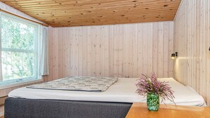 A bedroom with a wooden ceiling, a bed with a white mattress and a patterned blanket, a grey bench, a wooden nightstand with a vase of flowers, and a window with a view of trees. - 4 Person Holiday Home in Ebeltoft (Ebeltoft)