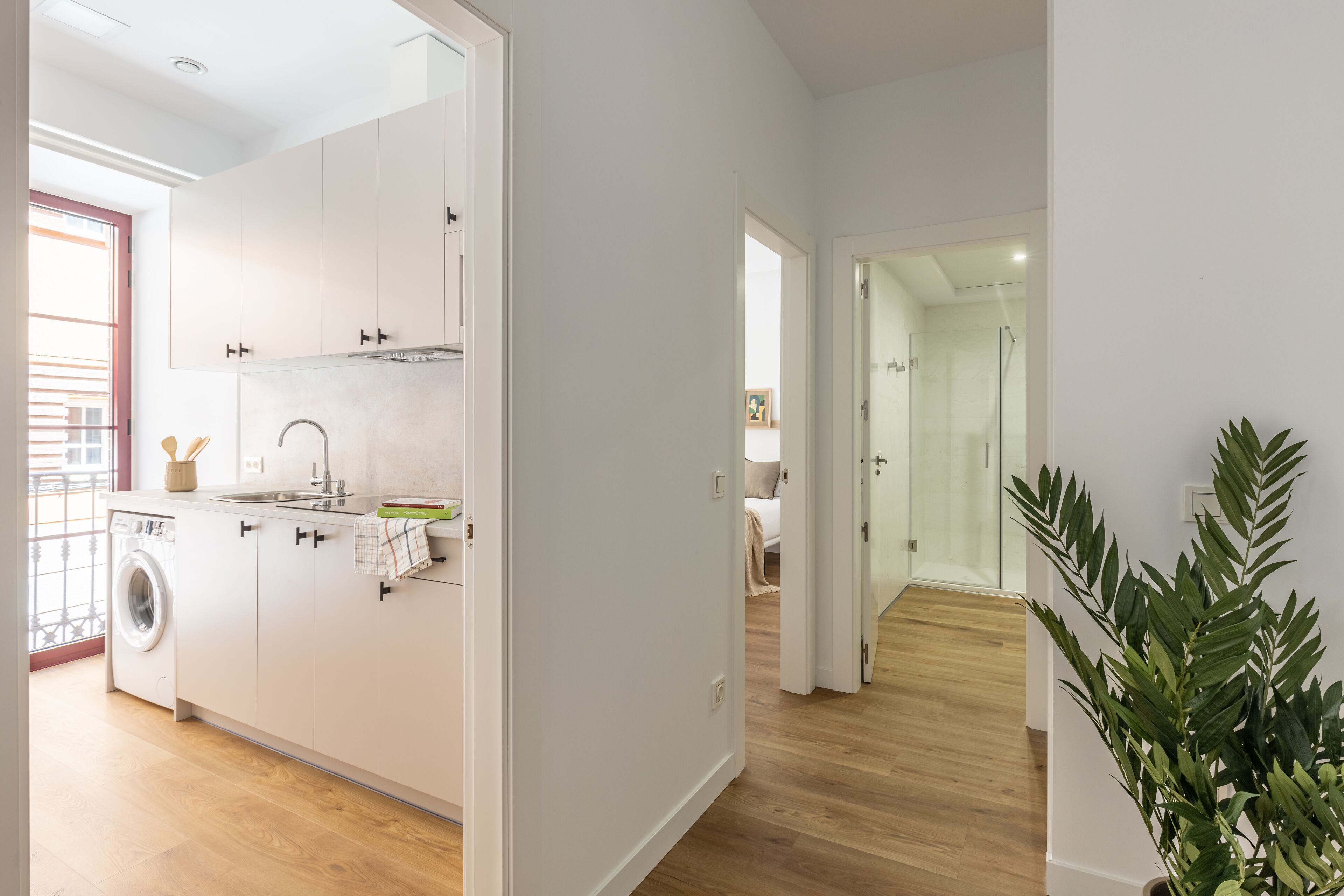 A modern kitchen with white cabinets, a washing machine, and a sink. There is a hallway leading to a bedroom with a glass door.