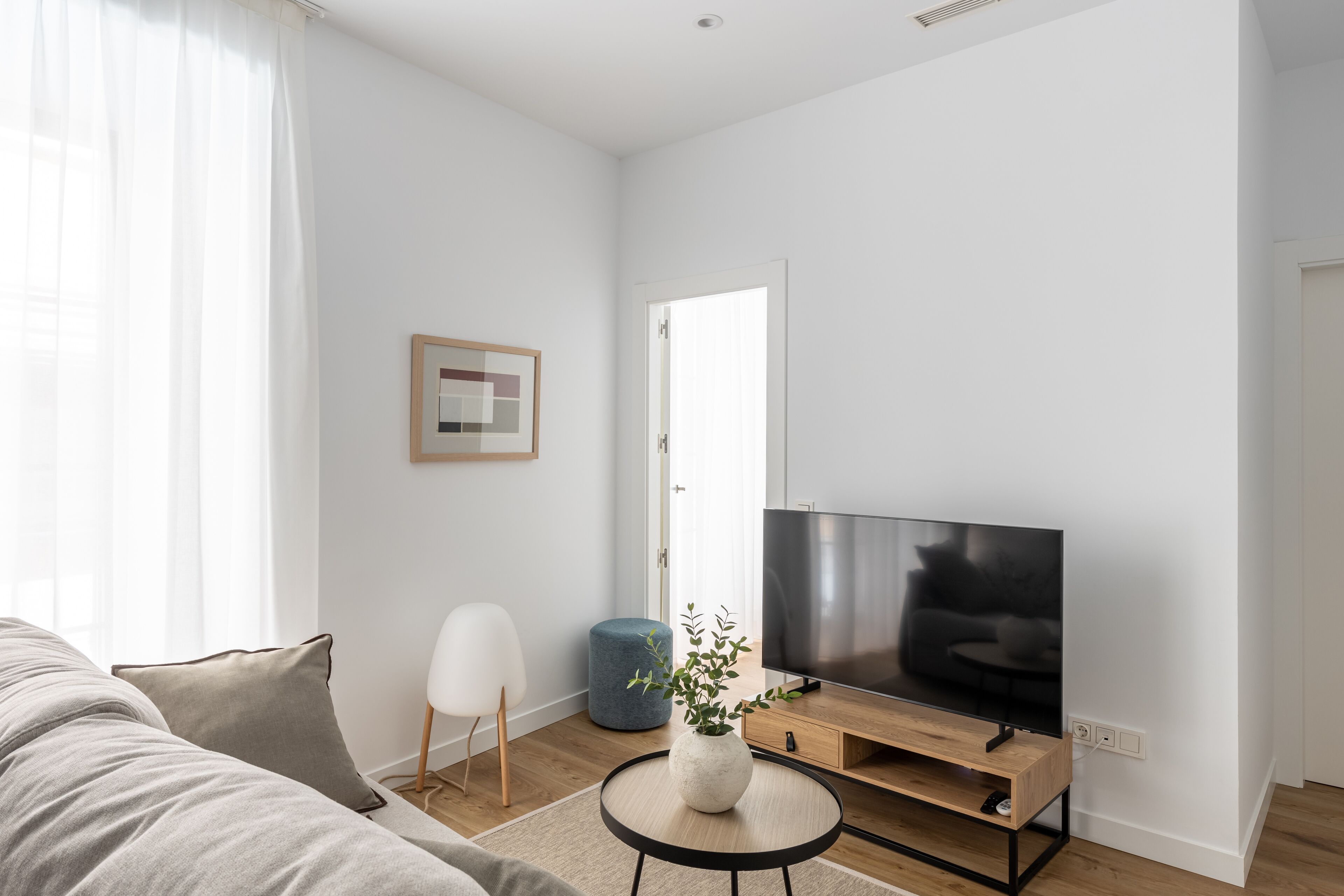 Ein modernes Wohnzimmer mit einem großen Flachbildfernseher, einem runden Couchtisch mit Pflanze, einem blauen Hocker und einem grauen Sofa.
