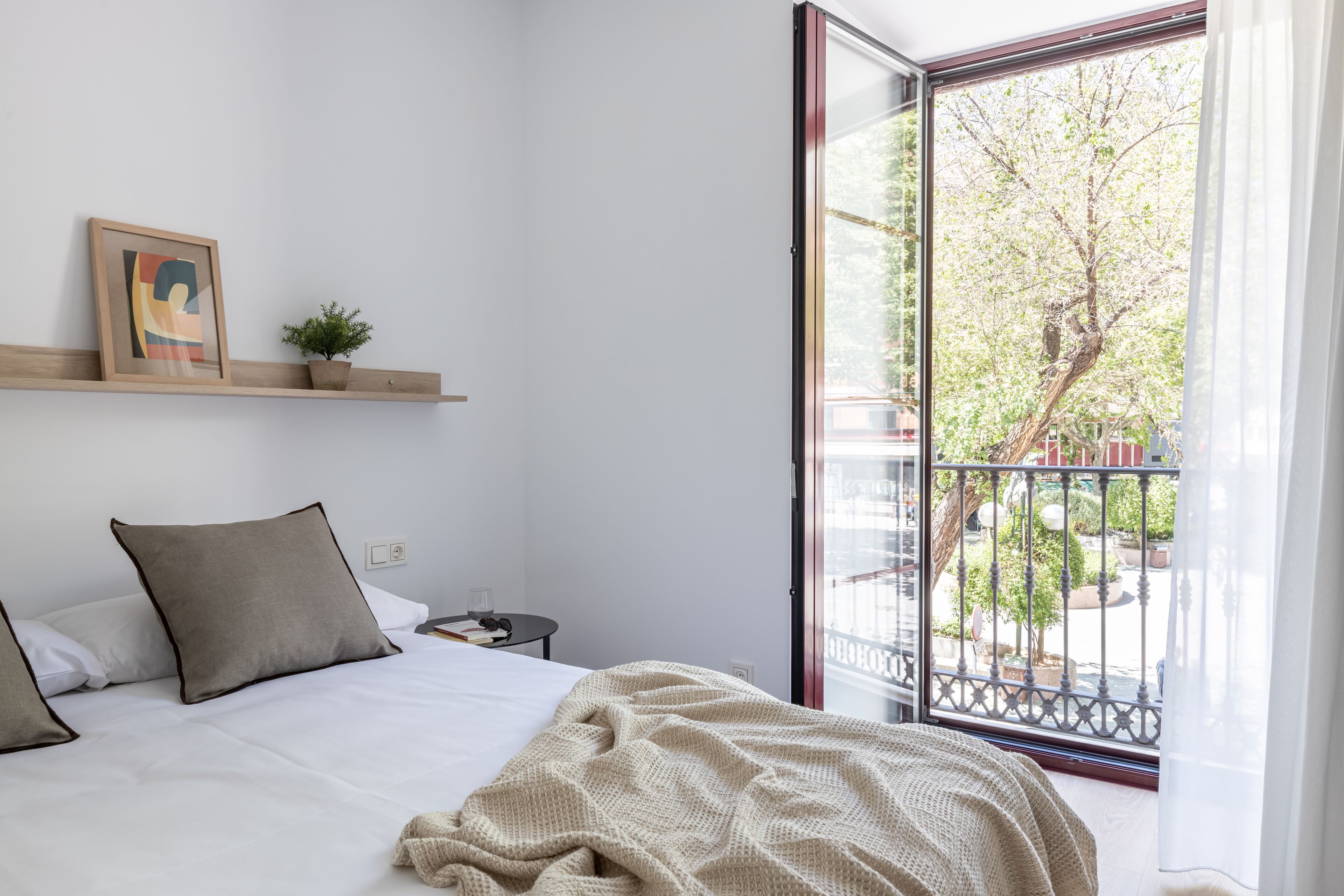 A bedroom with a bed, pillows, a bedside table, a small plant, a framed picture, and a balcony with a railing.