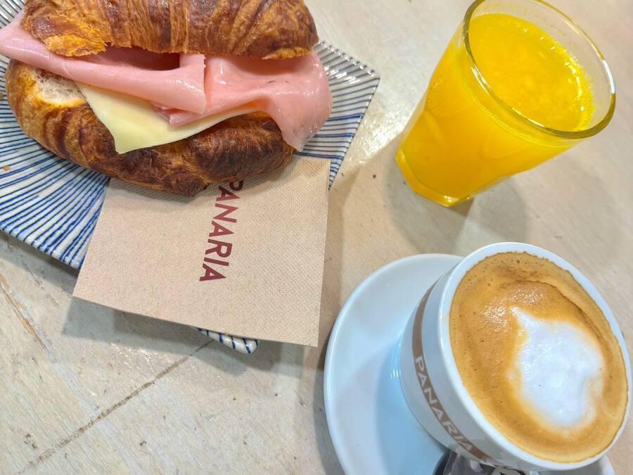 Un sándwich de croissant con jamón y queso, un vaso de jugo de naranja y una taza de café con arte de latte sobre una mesa.