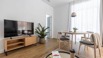 Ein modernes Wohnzimmer mit Flachbildfernseher, Holzmöbeln, einem Esstisch mit Stühlen und einem großen Fenster mit durchscheinenden Vorhängen.