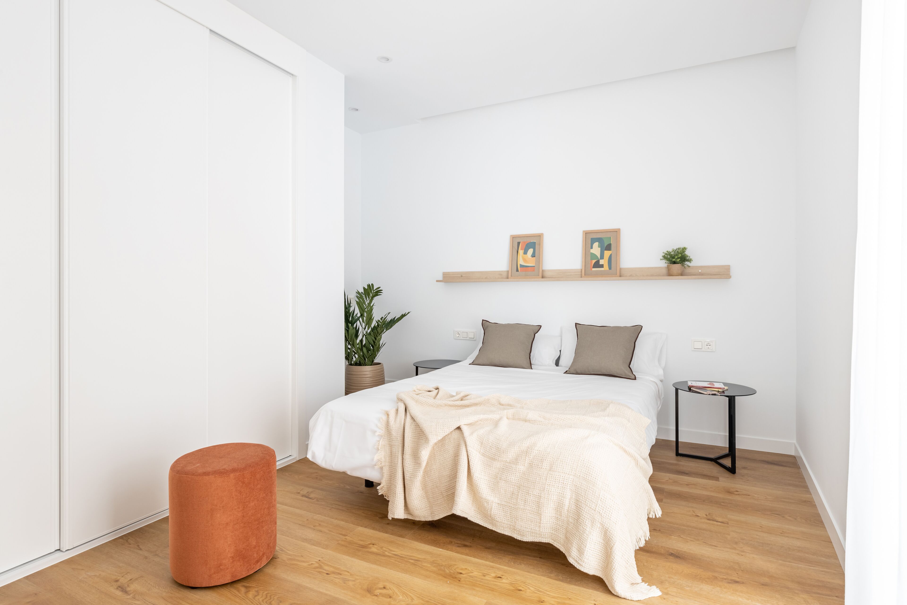 Un dormitorio minimalista con cama, una mesita auxiliar, una planta en maceta y un reposapiés rojo.