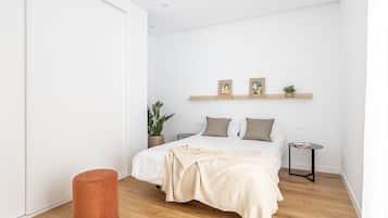 Un dormitorio minimalista con cama, una mesita auxiliar, una planta en maceta y un reposapiés rojo.