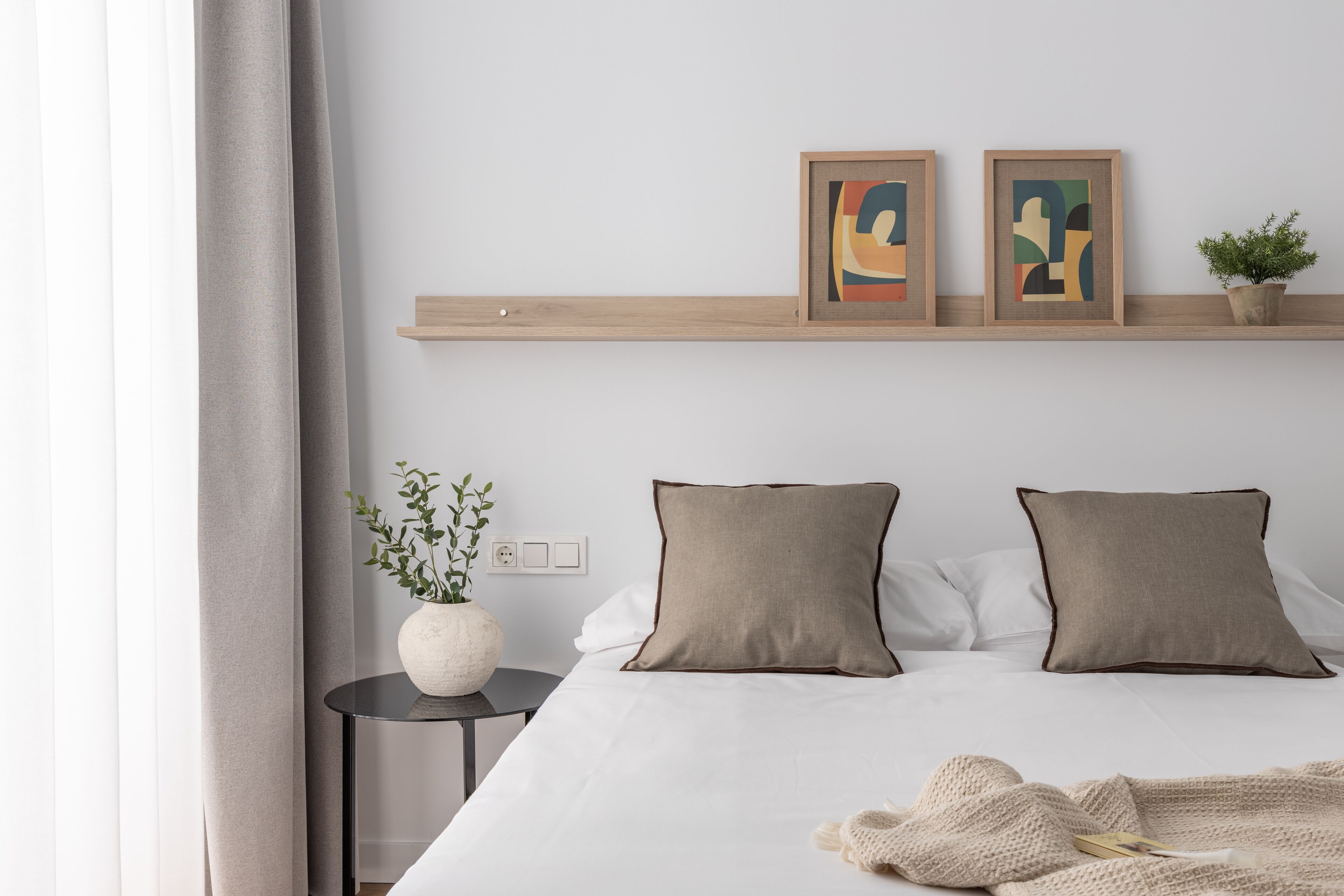 A neatly arranged bedroom with a bed, pillows, a small side table with a plant, and two framed artworks on a shelf above the bed.