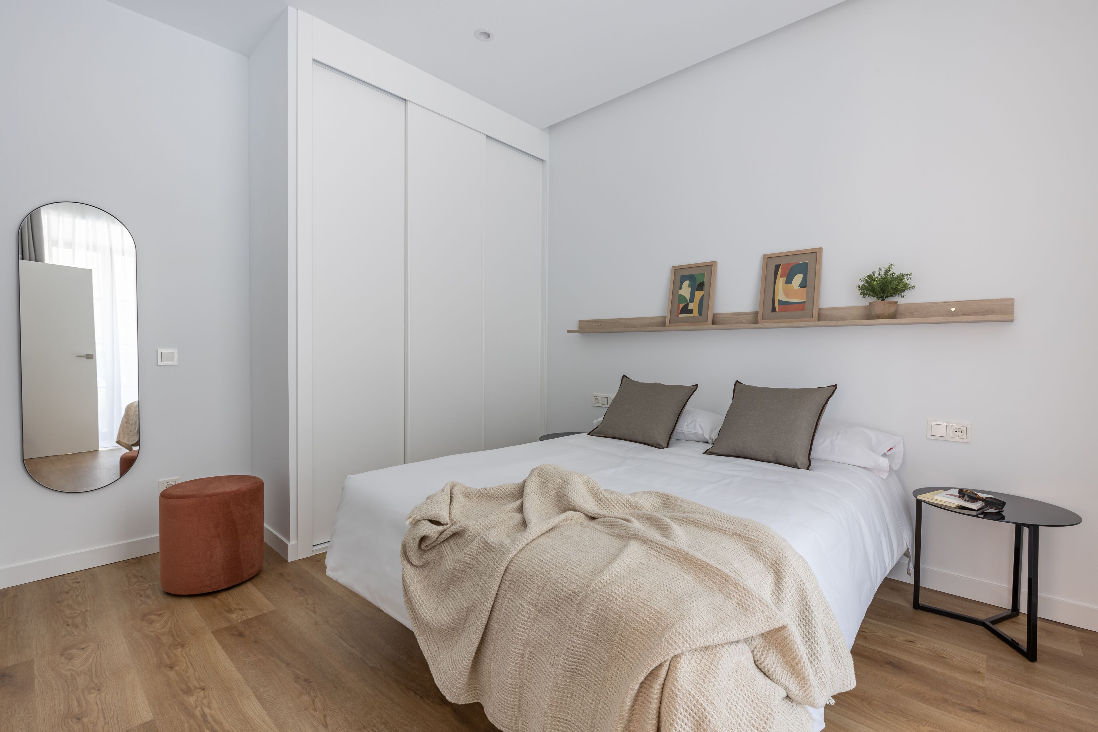 A modern bedroom with a bed, a round ottoman, a bedside table, and a wall-mounted shelf with pictures and a plant.
