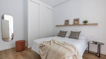 A modern bedroom with a bed, a round ottoman, a bedside table, and a wall-mounted shelf with pictures and a plant.
