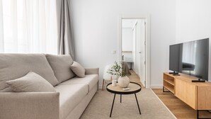 A modern living room with a sofa, a TV on a wooden stand, a small table with a vase, and a door leading to another room.