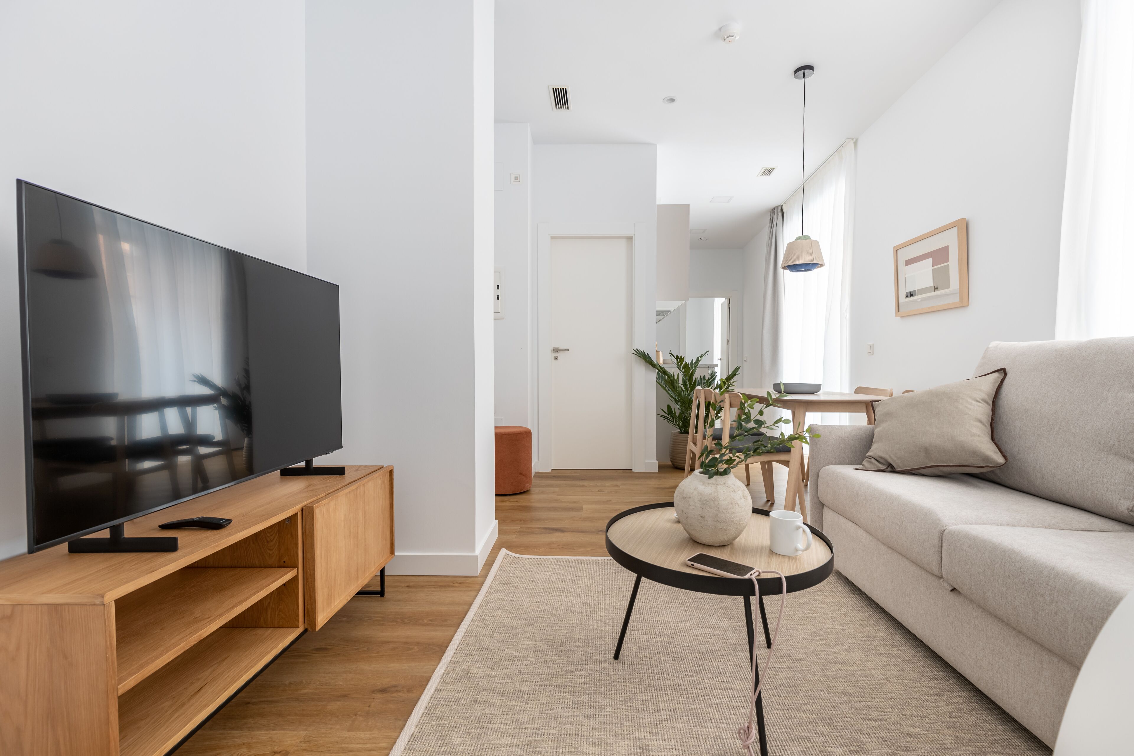 Een moderne woonkamer met een flatscreen televisie op een houten standaard, een ronde salontafel met een vaas en een afstandsbediening, een beige bank en een houten vloer.