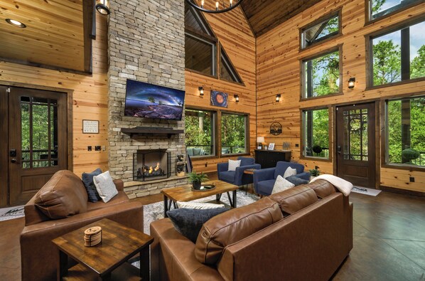 A spacious living room with a stone fireplace, leather sofas, a flat-screen TV, and large windows offering views of the surrounding forest.