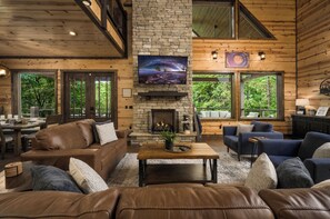 L’intérieur chaleureux d’un chalet avec une cheminée en pierre, des canapés en cuir et de grandes fenêtres donnant sur la forêt.