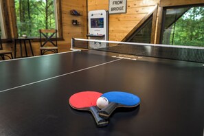 Una mesa de ping pong con dos paletas y una pelota, una pared de madera con un letrero y una ventana con vista a árboles.