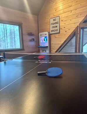 Una mesa de ping pong, una raqueta, un cartel que dice 'NO FISHING FROM BRIDGE' y una máquina de videojuegos en una habitación con paredes de madera y una ventana.