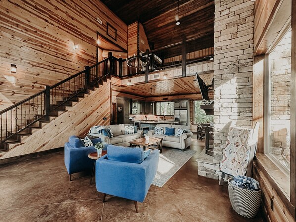 A spacious living area with a high ceiling, a staircase, and a stone fireplace. - Majestic Cabin Escape - 4 Bdr I Hot Tub I Fire Pit I Slide I Lake close by (Broken Bow)