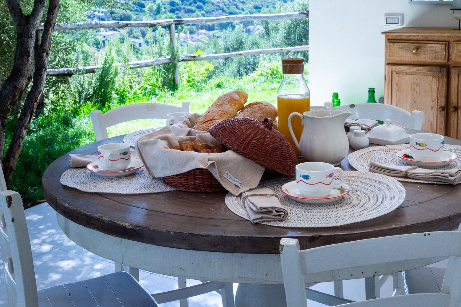 Une table ronde en bois, dressée pour le petit-déjeuner, avec du pain, une carafe, des tasses et une bouteille, et une vue panoramique en arrière-plan.