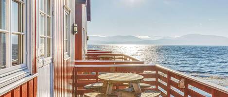 Una terraza de madera con una mesa redonda y sillas, con vista a una masa de agua y montañas al fondo.