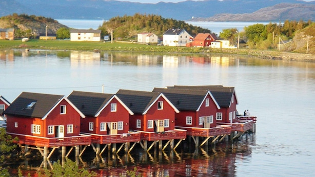Une rangée de maisons en bois rouge, construites sur pilotis au-dessus de l’eau, avec un lac paisible et des montagnes en arrière-plan.