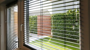 Een raam met horizontale zonwering, een witte wasbak en uitzicht op een bakstenen muur en begroeiing.