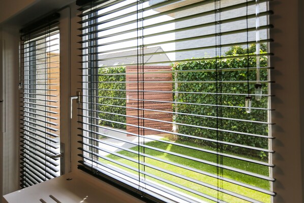 A window with horizontal blinds, a white sink, and a view of a brick wall and greenery outside.