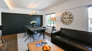 Een moderne woonkamer met een eethoek, met een grote wandklok, een zwarte leren bank en een houten salontafel met sinaasappels.