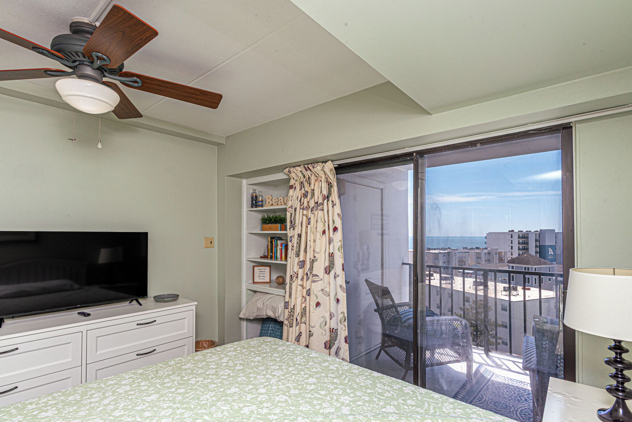 Un dormitorio con cama, televisión, ventilador de techo, balcón con sillas y vistas a edificios.