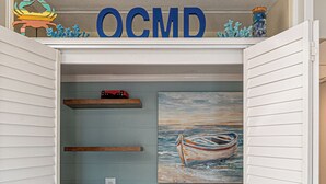 A laundry room with two washing machines, a shelf with decorative items, and a painting of a boat. - Bluewater -3 Bedroom Condo with Outdoor Pool! (Ocean City)