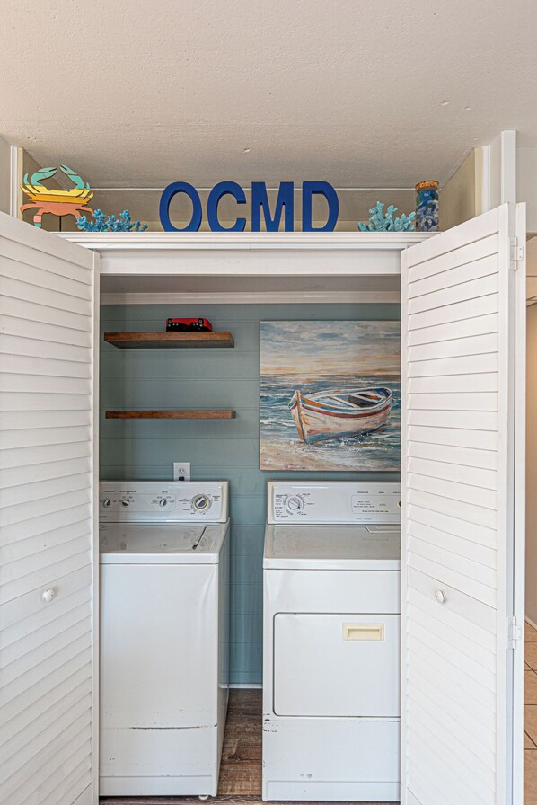 Une buanderie équipée de deux machines à laver, d’une étagère ornée d’objets décoratifs et d’un tableau représentant un bateau.