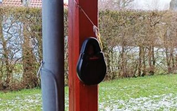 A red pole with a black object attached to it, a grey pipe, and a hedge in the background.