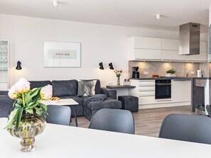 Un salon moderne avec un canapé gris, une table à manger avec des chaises et un coin cuisine doté de meubles blancs et d’une hotte de cuisinière en acier inoxydable.