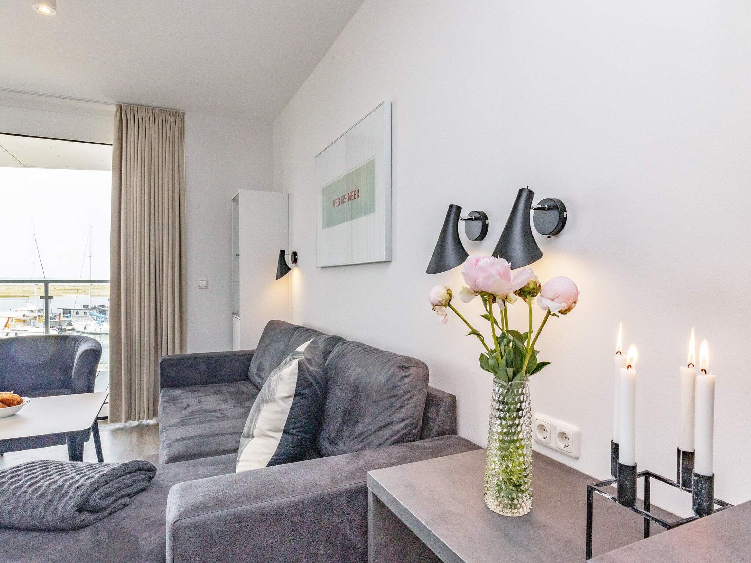 A modern living room with a grey sofa, a small table with a vase of flowers, and two wall-mounted lamps.