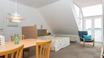 A room with a sloped ceiling, a bed with a patterned duvet, a wooden dining table with chairs, a blue chair with a cushion, and a hanging light fixture.