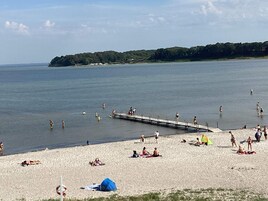 Una spiaggia con persone che nuotano e si prendono il sole, un molo che si protende nell'acqua e un'isola boscosa all'orizzonte.